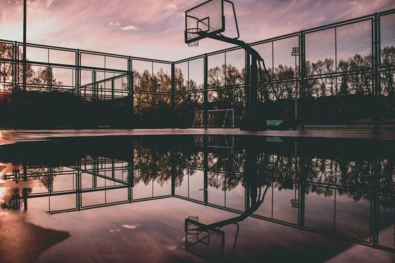 A serene sunset casts reflections on a wet basketball court, creating a tranquil urban scene.