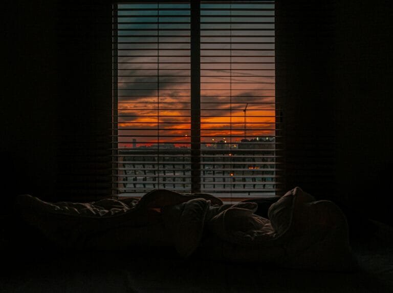 Cozy bedroom with a view of a vibrant sunset through window blinds.
